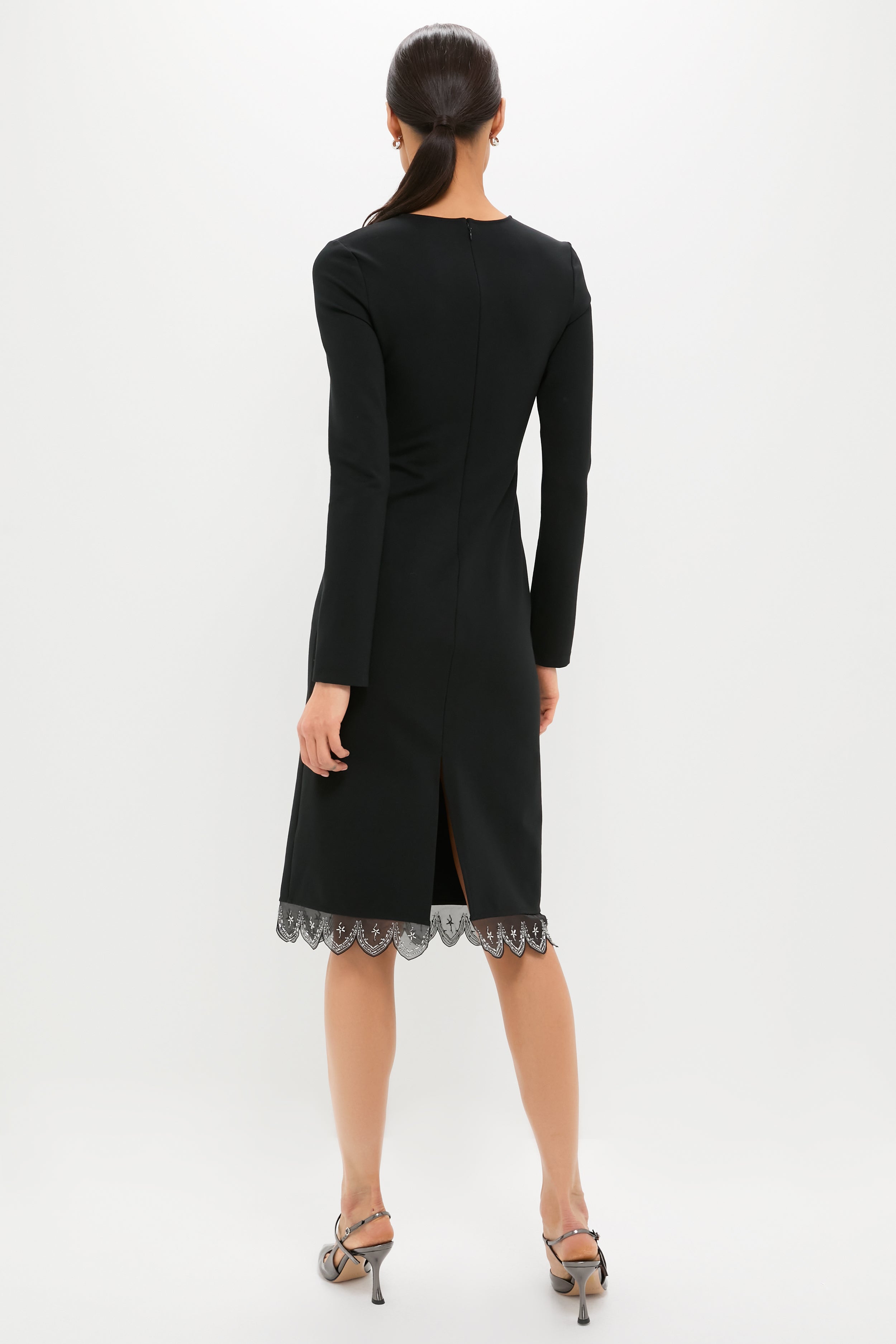 Woman in a classic Black Hunter Dress with scalloped lace hem, effortlessly styled with metallic-grey sandals, embodying high-end, fun elegance against a simple backdrop.