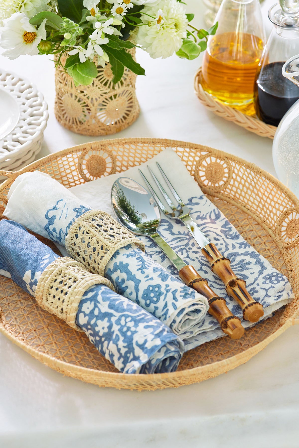 Effortlessly styled Raffia Round Tray with blue-patterned napkins, bamboo-handled cutlery, and blooms, embodying a classic, high-end coastal design for a fun, versatile tabletop display.