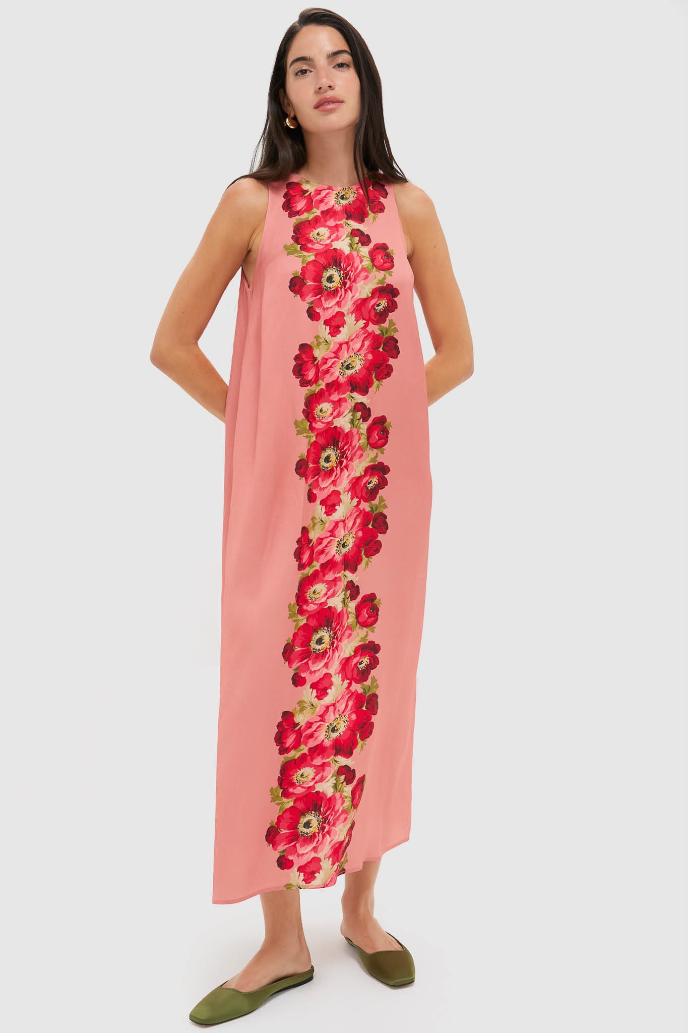 A woman wears a Tuckernuck sleeveless, ankle-length pink floral maxi dress with a vertical row of red and green patterns down the center, paired with green flat shoes.