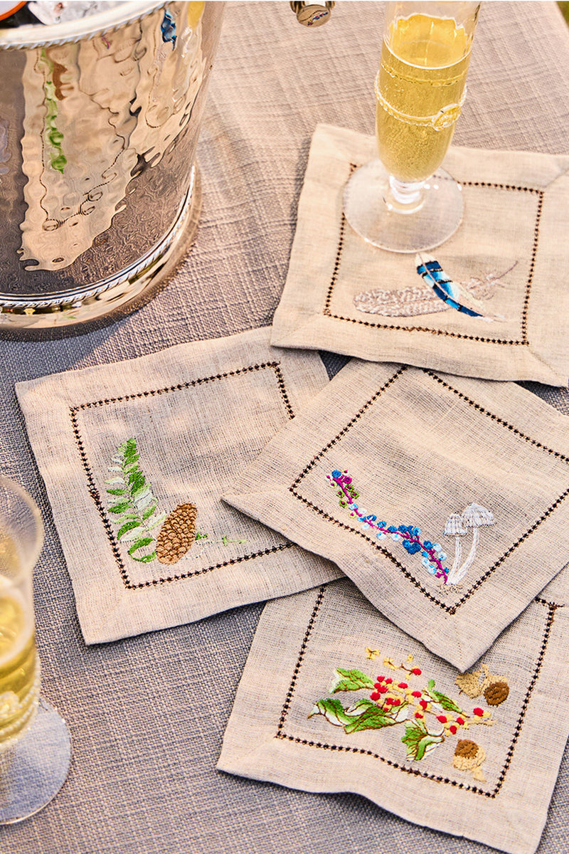 Forest Walk Cocktail Napkin Assorted Set of 4, featuring hand-embroidered woodland motifs, elegantly arranged with champagne flutes and an ice bucket, epitomizing classic, effortless, high-end table decor.