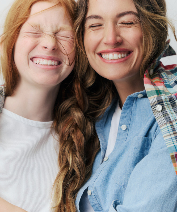 Close up of two people smiling. The girl to the left is wearing a white tee. The woman to the right is wearing a white tee layered with a chambray and a madras shirt. 