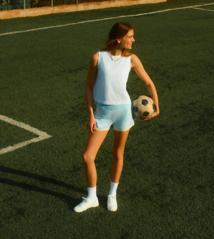Woman standing on a soccer field holding a soccer ball in a blue tank and shorts 