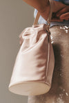 Blush Lila Mini Bag: A classic, pale-pink satin bucket bag with leather strap, effortlessly held by a hand, perfect for high-end, fun evenings.