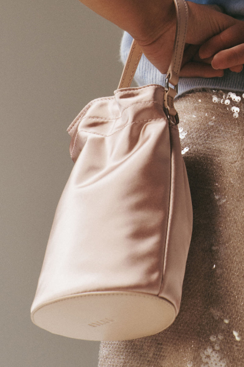 Blush Lila Mini Bag: A classic, pale-pink satin bucket bag with leather strap, effortlessly held by a hand, perfect for high-end, fun evenings.