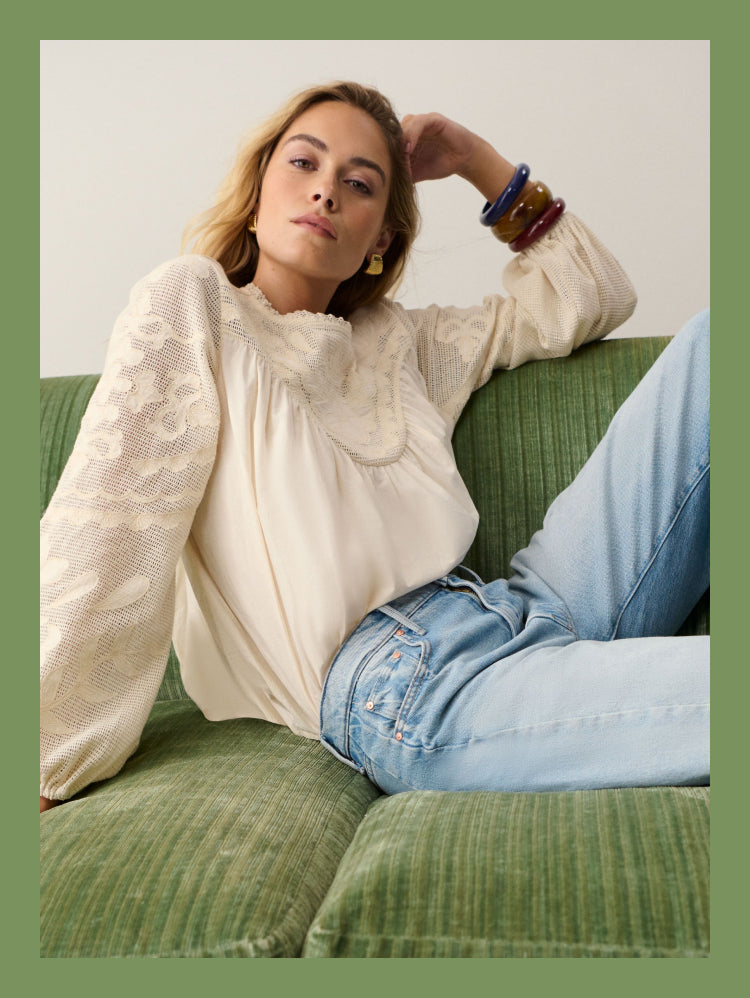 Woman sitting on a green couch in a white long sleeved shirt and light wash jeans. 