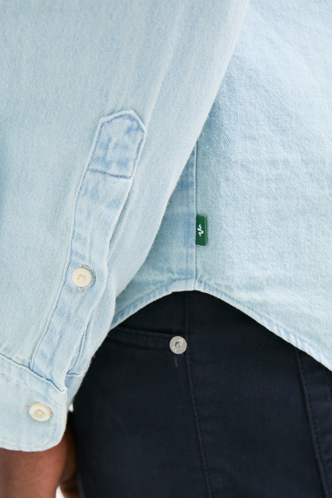 Classic Light Blue Washed Denim Shirt drapes effortlessly over dark-navy pants, showcasing a stylish, high-end look with button cuffs and a green brand tag.