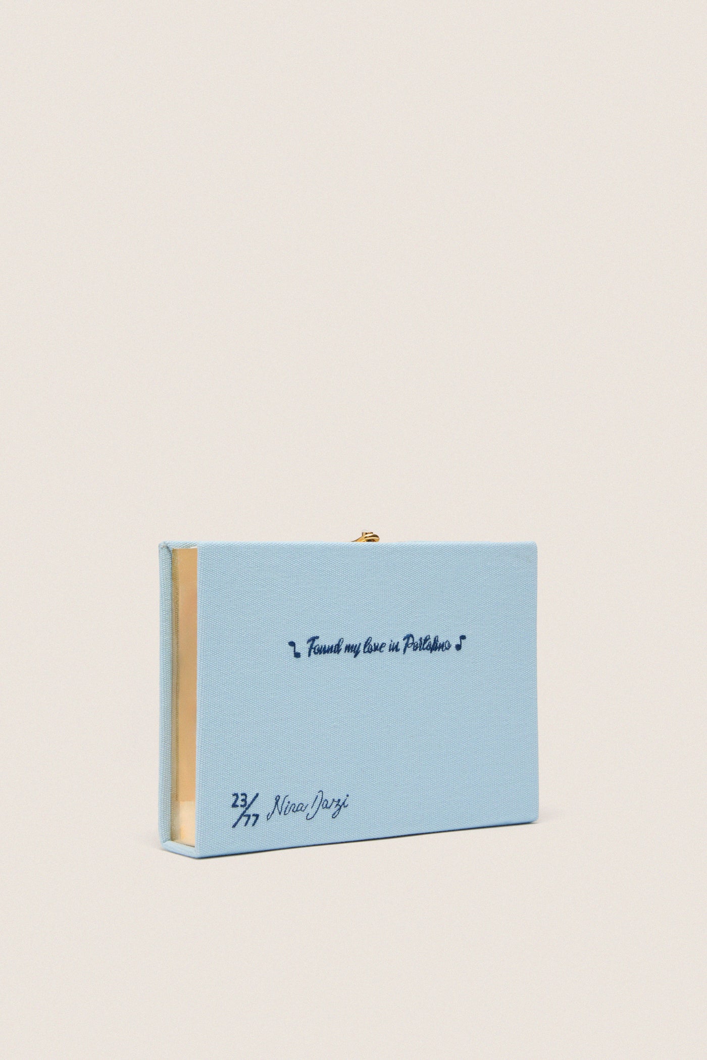 Portofino Book Clutch, a limited-edition, pastel-blue embroidered accessory with gold trim, combines classic elegance and high-end craftsmanship, embodying effortless style and playful sophistication.