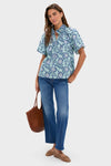 Woman models the classic, effortless Multi Blue Leafy Floral Thea Blouse with keyhole neckline, paired with mid-rise jeans, taupe ballet flats, and woven straw tote.