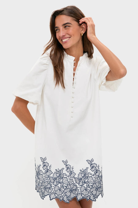 Smiling woman in the White & Navy Embroidery Mimi Mini Dress showcasing classic, effortless style with puff sleeves, ruffle collar, and embroidered hem, embodying fun, high-end fashion.