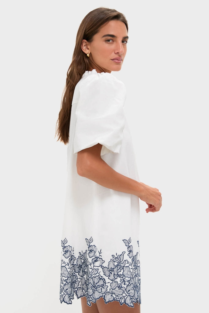 Model in profile wears the White & Navy Embroidery Mimi Mini Dress, showcasing classic, effortless style with floral embroidery, ruffle collar, and puff sleeves for a fun, high-end look.