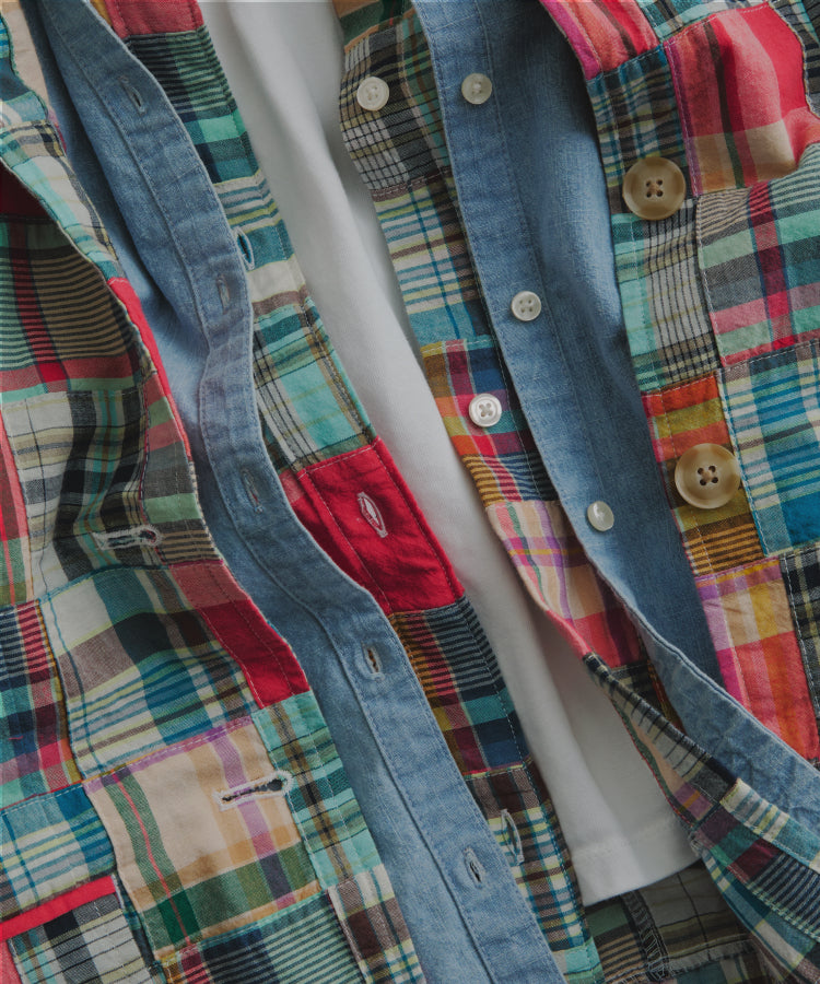 Several shirts layered together. The base is a white tee and then three shirts are layered on top of it. A madras shirt with white buttons, a chambray shirt with white buttons, and a madras shirt with chambray buttons. 