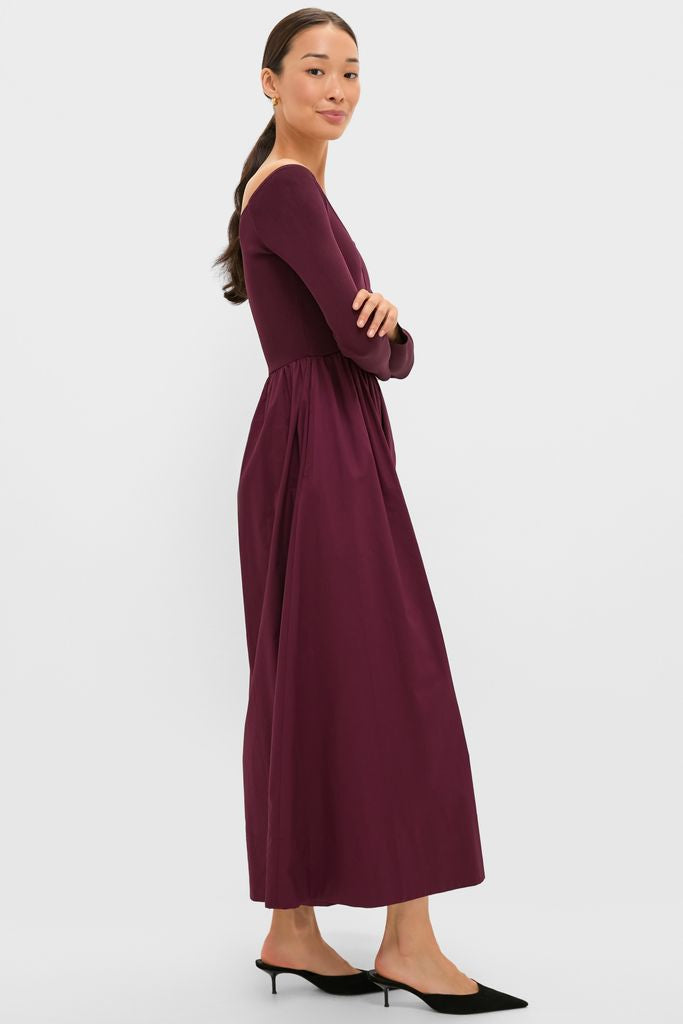 Woman wearing the Winetasting Clea Dress, a classic, high-end burgundy gown with long sleeves and a gathered skirt, styled with black mules and gold hoops.