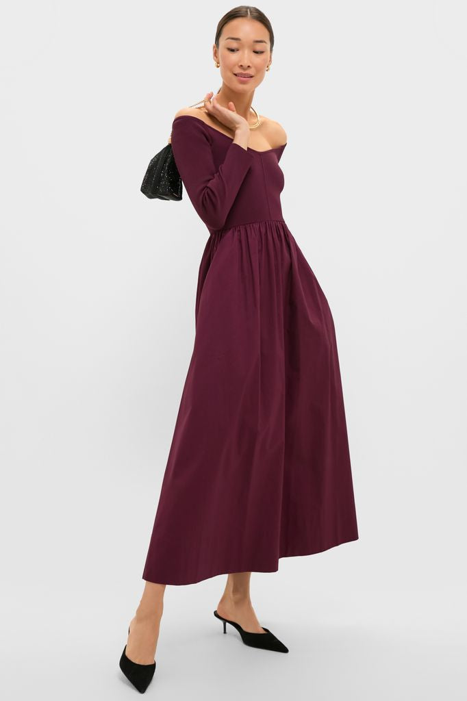 Woman wearing the classic Winetasting Clea Dress, an off-the-shoulder, fit-and-flare gown, styled effortlessly with black mules, gold jewelry, and a chic handbag.