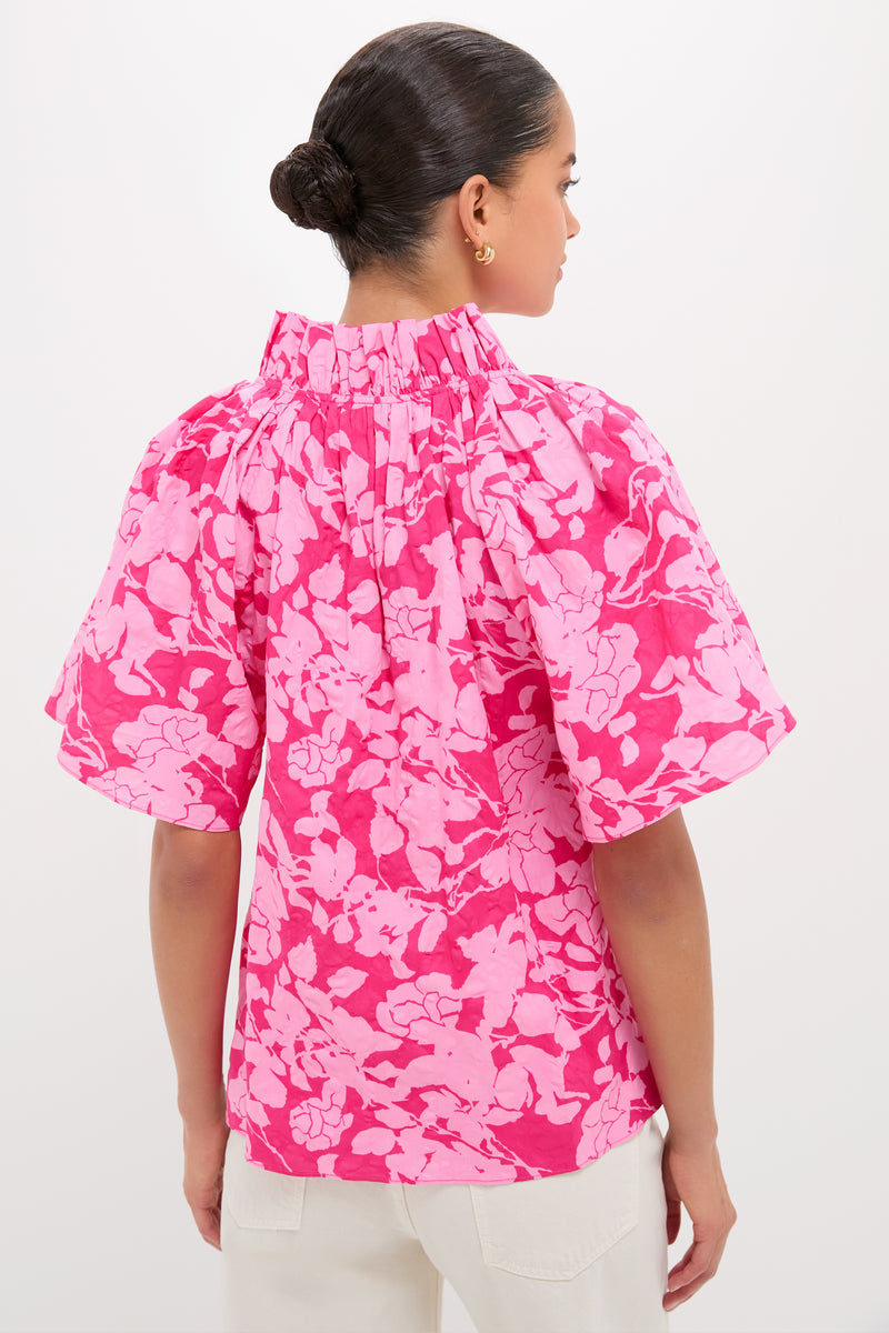 Back view of a model in the classic, high-end Pink Textured Floral Thea Blouse with ruffled collar and billowy sleeves, paired effortlessly with light pants.