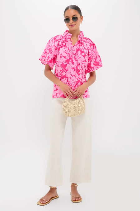 Woman in Pink Textured Floral Thea Blouse with ruffled neckline, paired with high-waisted ivory pants, gold sandals, and a straw handbag for an effortless, high-end look.