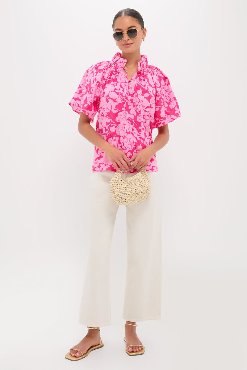 Woman in Pink Textured Floral Thea Blouse with ruffled neckline, paired with high-waisted ivory pants, gold sandals, and a straw handbag for an effortless, high-end look.