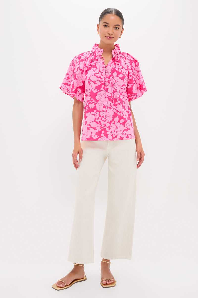 Woman models the Pink Textured Floral Thea Blouse, featuring a relaxed fit, ruffle collar, and flutter sleeves, paired effortlessly with high-waisted wide-leg pants and gold sandals.