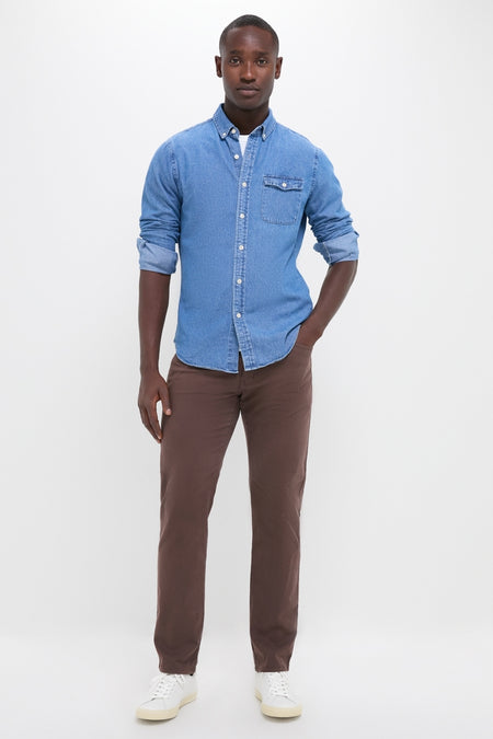 Man models Dark Indigo Washed Denim Shirt; a classic, effortless piece with button cuffs and tab collar, styled casually with brown pants and white sneakers.