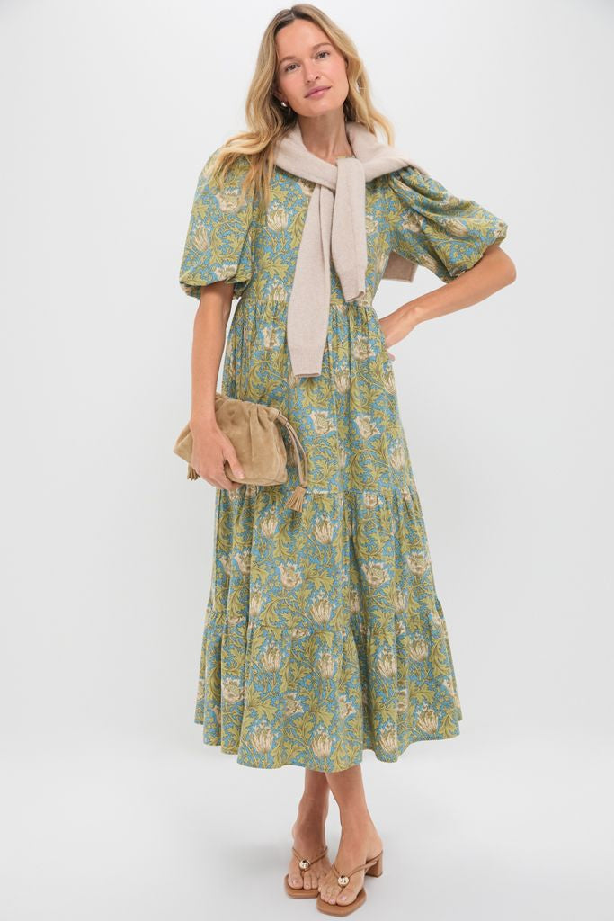 Woman in classic Blue & Green Leafy Floral Cisco Maxi Dress with puff sleeves, taupe clutch, and sandals, embodying effortless, fun, high-end style.
