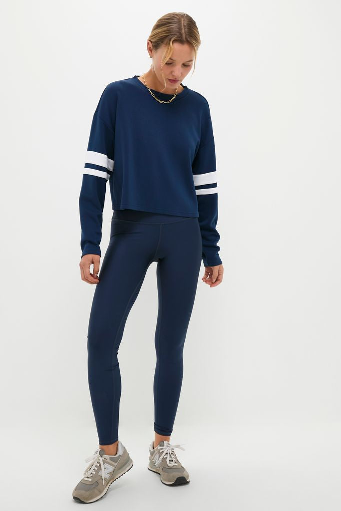 Woman wearing Navy Varsity Comfort Stretch Matilda Pullover with white arm stripes, paired with matching leggings and gray sneakers, showcasing a classic, effortless, and high-end athletic style.