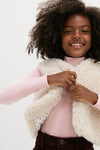 Young girl smiles while adjusting an effortlessly classic, high-end Cream Teddy Vest, perfect for fun, stylish layering, embodying Tuckernuck's chic, confident fashion ethos.