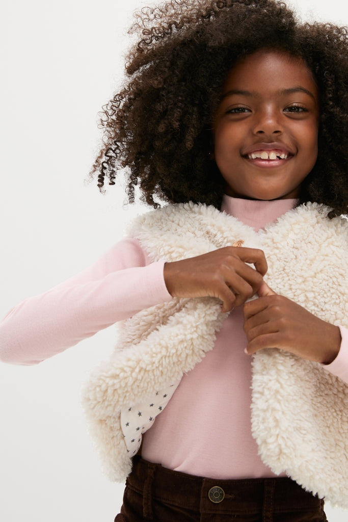 Young girl smiles while adjusting an effortlessly classic, high-end Cream Teddy Vest, perfect for fun, stylish layering, embodying Tuckernuck's chic, confident fashion ethos.
