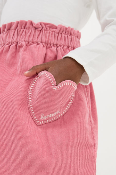 Child's hand in heart-pocket of classic Pink Douchka Skirt, showcasing effortless French style with fun, high-end details like bonjour embroidery and elastic waist.