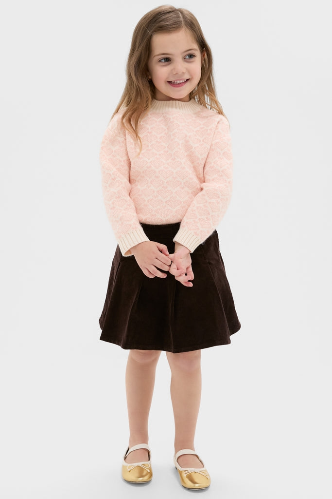 Young girl wearing a classic Light Pink Heart Sweater with ribbed knit finishes, paired effortlessly with a dark brown A-line skirt and gold Mary Jane shoes, exuding fun, high-end style.