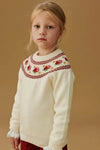 Ivory Grenada Fair Isle Merino Wool Girl Jumper with classic intarsia patterns, effortlessly modeled for a fun, high-end look, paired with deep red trousers.