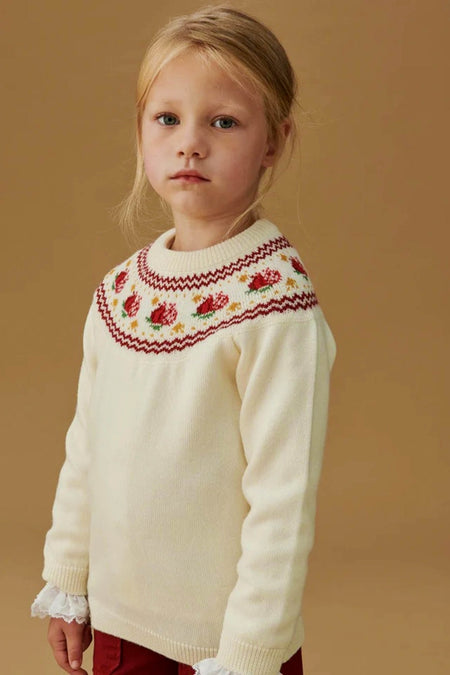 Ivory Grenada Fair Isle Merino Wool Girl Jumper with classic intarsia patterns, effortlessly modeled for a fun, high-end look, paired with deep red trousers.