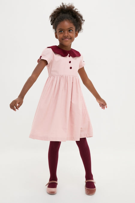 Young girl in classic, high-end Pink Julieta Girl Dress with a velvet burgundy collar, effortlessly playful in ribbed tights and Mary Jane shoes, embodying fun timeless style.