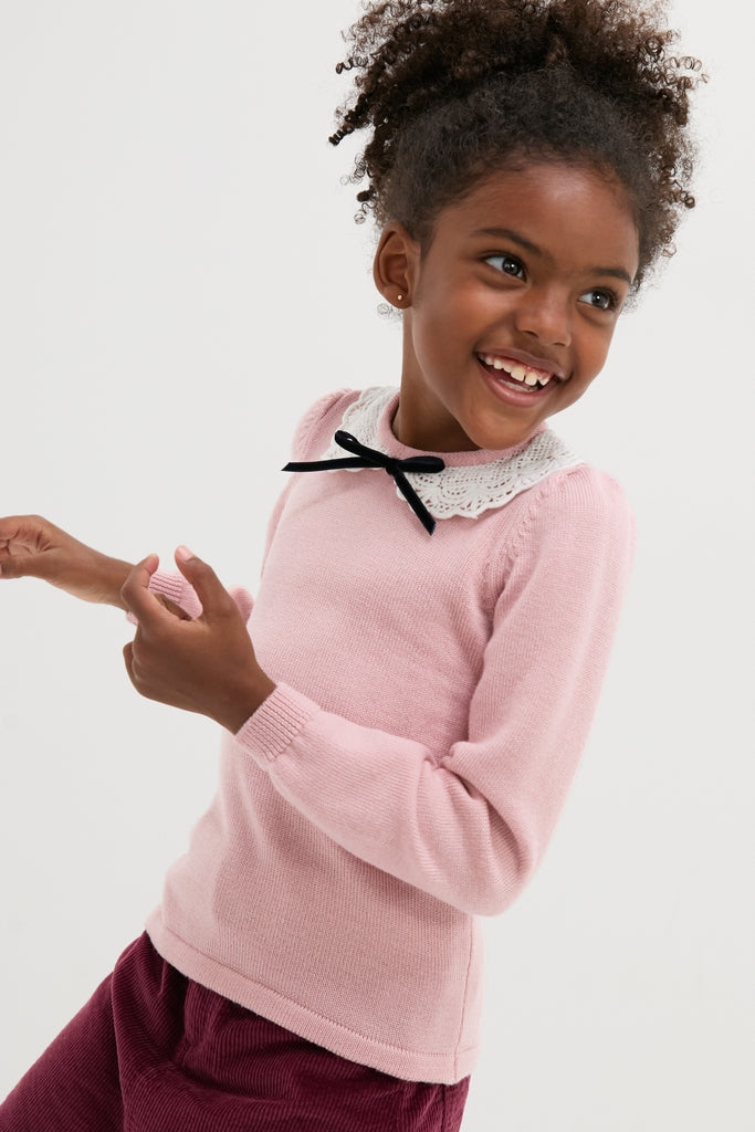 Young girl wearing the classic Dusty Pink Blanca Merino Wool Girl Jumper with lace collar and black bow, embodying effortless, high-end style and fun sophistication.