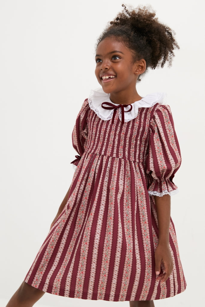 Young girl in a classic Burgundy Floral Stripe Festa Girl Hand-Smocked Dress, featuring an effortless fit-and-flare silhouette, lace trims, and a charming Peter Pan collar.