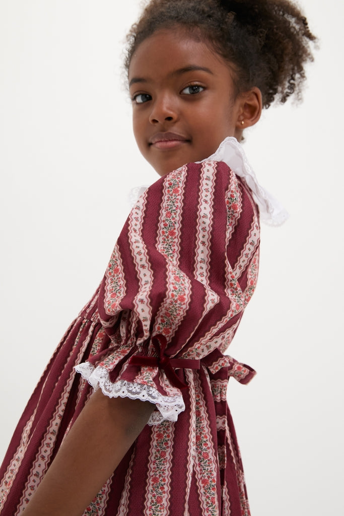 Young girl in a classic burgundy floral stripe Festa Girl Hand-Smocked Dress, showcasing effortless style with elbow-length puff sleeves and a charming lace-trimmed Peter Pan collar.