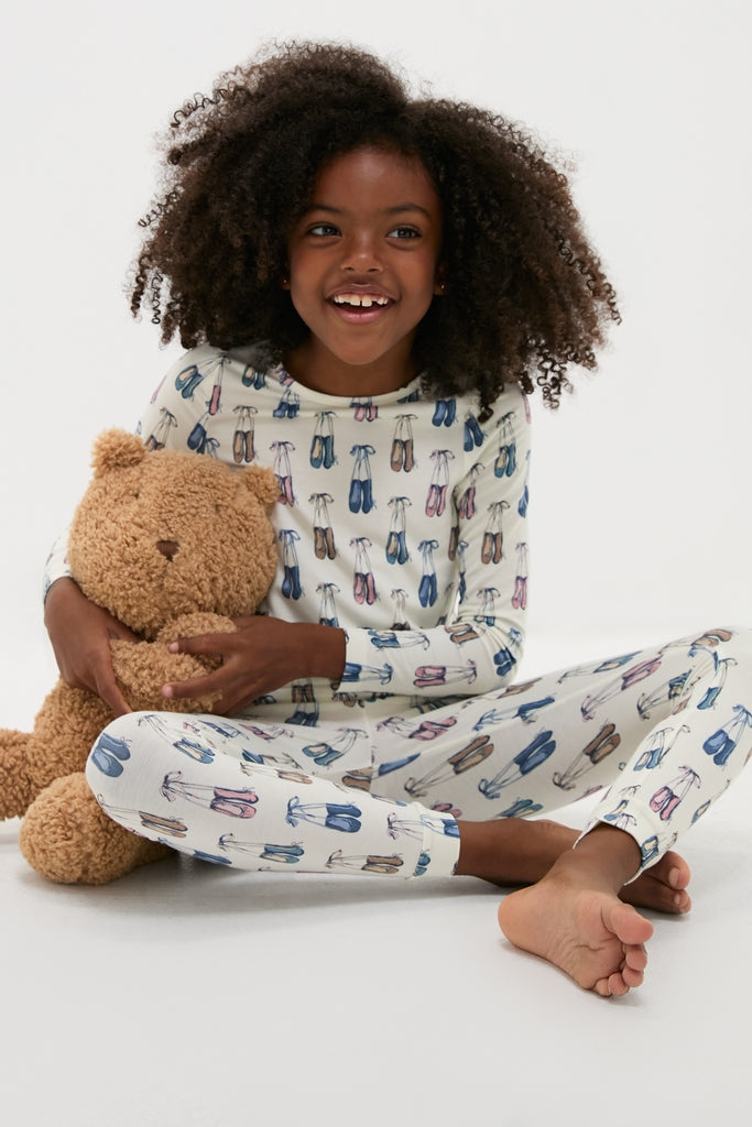 Young girl in the Ballet Slippers Kids Bamboo Pj Set, featuring an ice skate pattern, hugging a teddy bear. The classic, effortless pajama set is both fun and high-end.