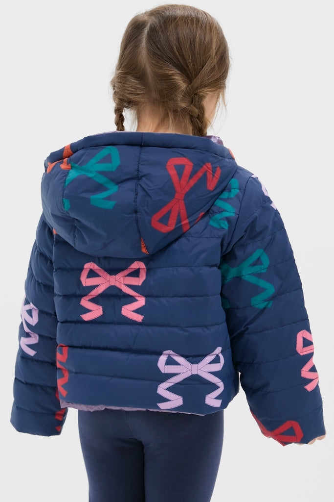 Young girl in a classic, fun, navy-blue Girls Reversible Riley Jacket with colorful bows, embodying effortless style and high-end versatility against a light background.