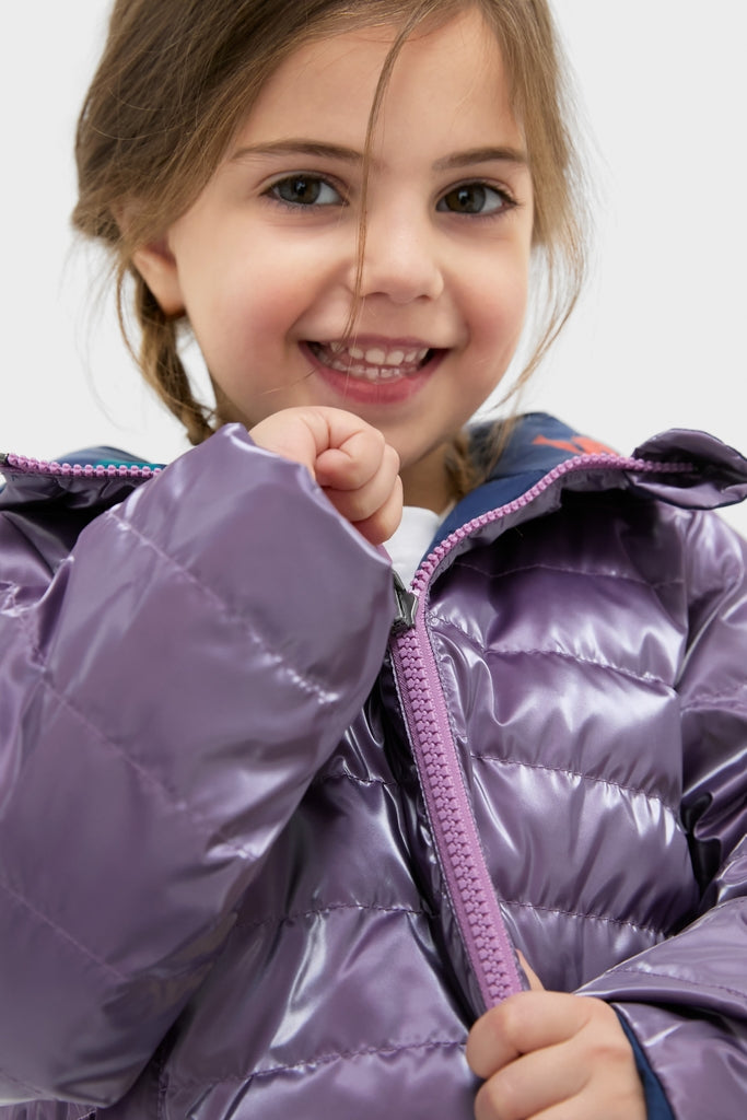 Young girl beams while zipping the Girls Reversible Riley Jacket, showcasing its classic, fun, and effortless high-end design with a metallic purple side.