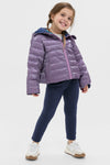 Young girl models the classic Girls Reversible Riley Jacket, showcasing its effortless, fun, high-end style. The jacket's metallic purple side is visible, complemented by navy leggings and white sneakers.