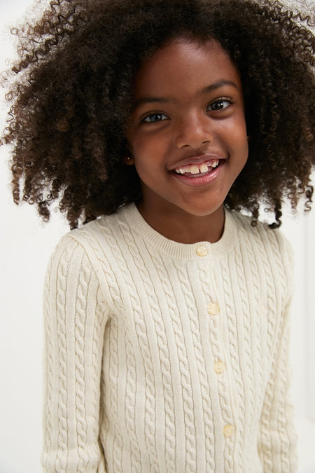 Young girl smiles, wearing Polo Ralph Lauren's Girls Long-Sleeve Cotton Cable Knit Lurex Cardigan, showcasing a classic, high-end, and effortlessly stylish design with button placket and ribbed hems.