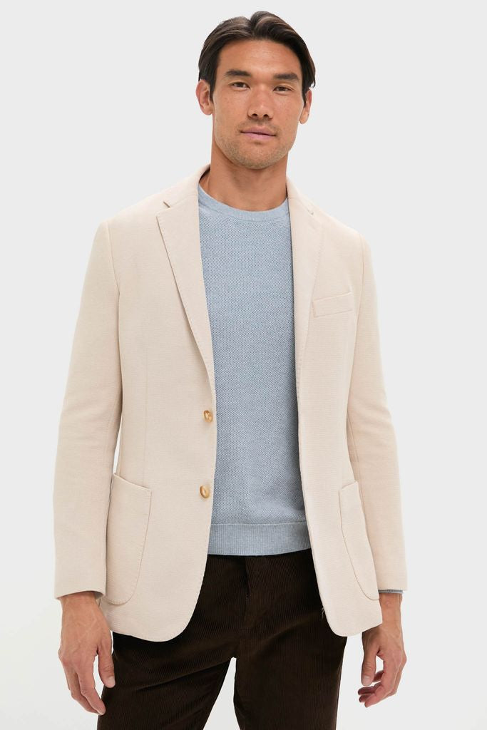 Man wearing the classic Tan Knit Travel Blazer by Tuckernuck, styled with a pale blue sweater and brown trousers, embodying effortless, high-end travel readiness.