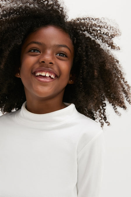 Young girl with curly hair wearing the White Frank T-Neck, a classic, effortless, high-end turtleneck from Oso & Me, showcasing its soft, breathable organic cotton fabric.