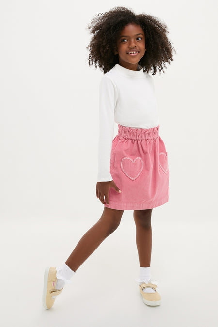 Young girl in a classic White Frank T-Neck, effortlessly styled with a pink skirt featuring heart pockets, embodying a fun and high-end, confident look.