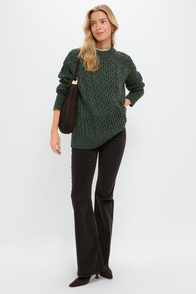 Young woman models the Hunter Green Galway Fisherman Sweater, showcasing its classic cable-knit design with a relaxed fit, paired effortlessly with dark flared trousers and suede accessories.