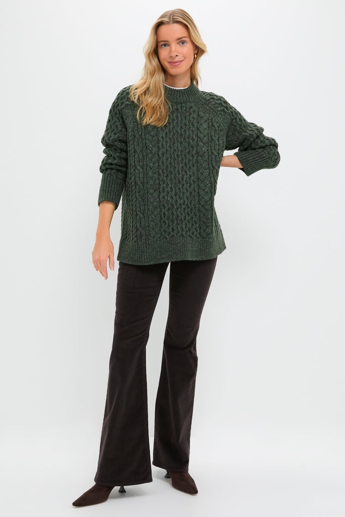 Person in chunky, forest-green Hunter Green Galway Fisherman Sweater, showcasing classic cable-knit design and relaxed fit, paired with dark flared pants and pointed-toe shoes for effortless style.