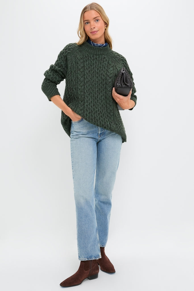 Model wearing the Hunter Green Galway Fisherman Sweater, a classic, oversized cable-knit with ribbed trim, styled effortlessly with jeans and a quilted handbag for a high-end, fun look.