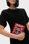 Person holding Bordeaux The Nutcracker Book Clutch, featuring an embroidered nutcracker and ornaments. Paired with simple gold jewelry, it’s a classic, high-end accessory for book lovers.