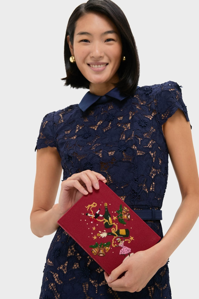 Young woman holds Camelia Holly Jingle and Mingle Book Clutch, embodying classic, effortless elegance. Features festive motifs, perfect for book lovers and high-end fashion enthusiasts.
