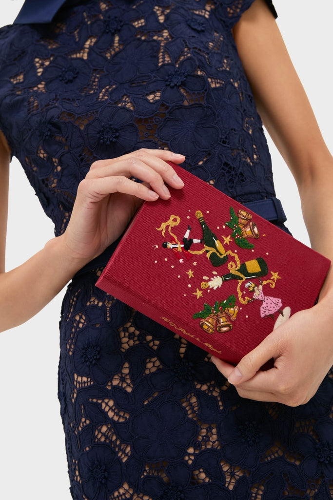 Person holds the classic Camelia Holly Jingle and Mingle Book Clutch, featuring festive embroidery, embodying high-end, effortless style and fun for book lovers.