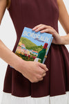Person wearing a sleeveless burgundy dress holds the Hortensia Bordeaux Book Clutch, a classic, high-end accessory with a fun, embroidered French townscape design. Effortlessly cool and unique.