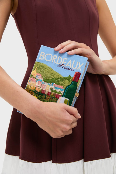 Person wearing a sleeveless burgundy dress holds the Hortensia Bordeaux Book Clutch, a classic, high-end accessory with a fun, embroidered French townscape design. Effortlessly cool and unique.